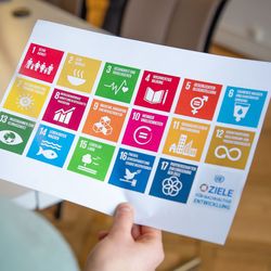 Eine Hand hält ein Blatt mit einer Abbildung der SDGs, farbige Kacheln, hoch.